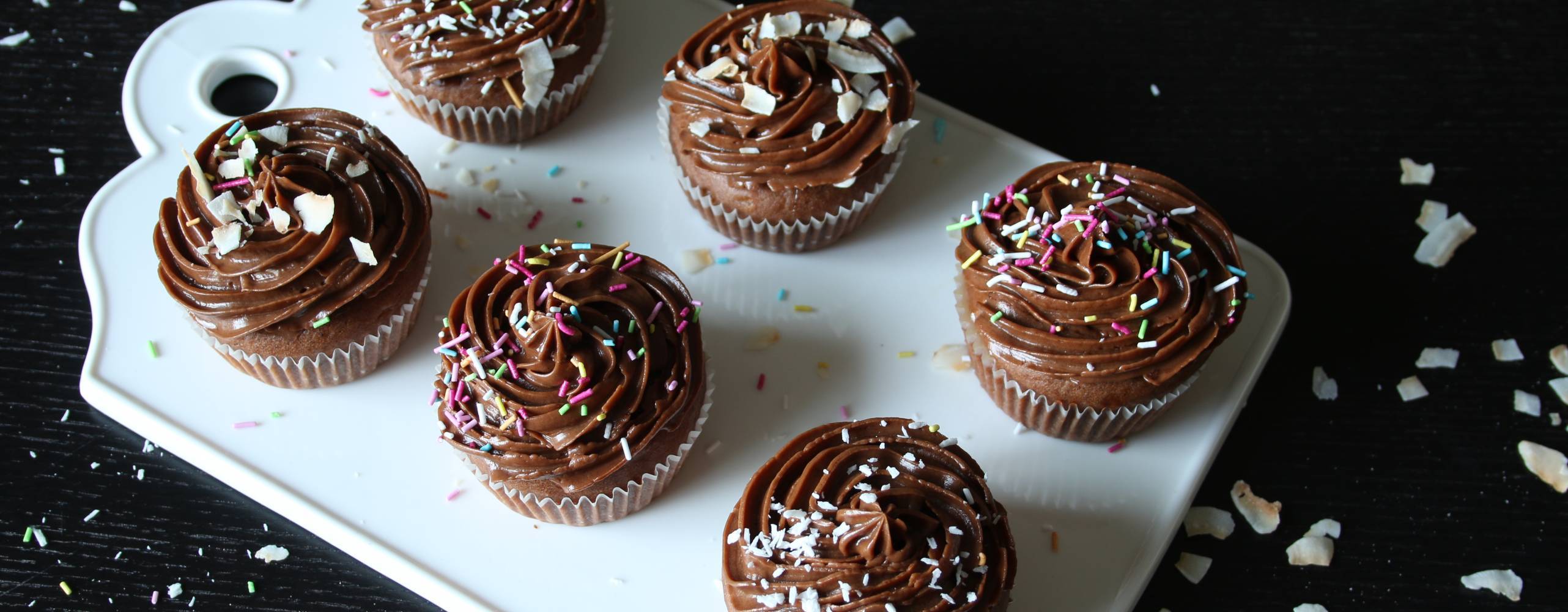 Sex stycken kolamuffins med mockaglasyr, kokos och strössel på en vit skärbräda på ett svart bord.