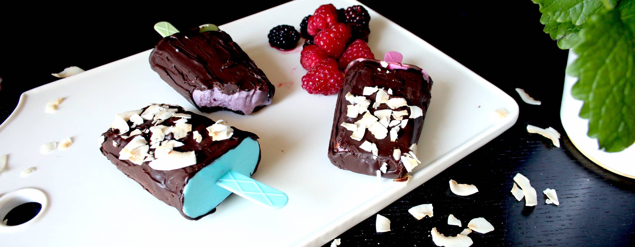 Tre chokladdoppade soygurtglassar på en vit skärbräda, bredvid glassarna ligger några hallon och björnbär