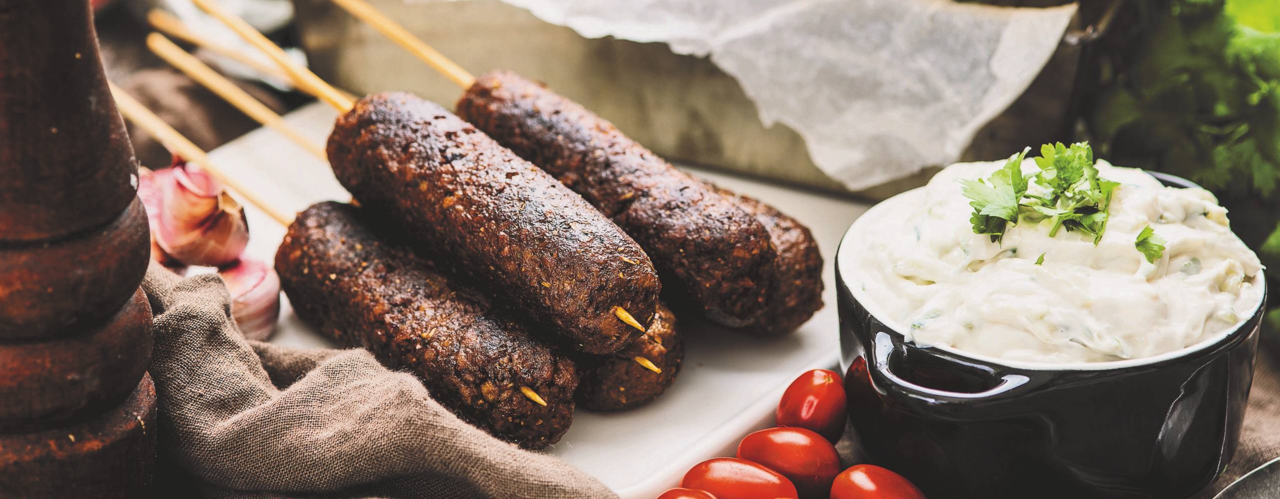 Kebabspett på formbar färs kebabspett på en vit skärbräda, bredvid spetten står en skål med tzatziki