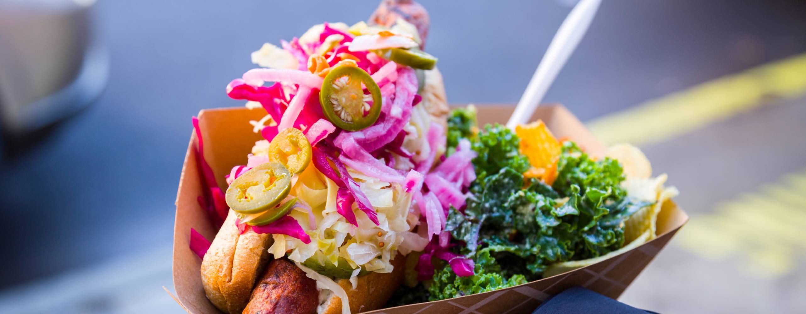 Hand håller upp bröd med vegansk fyllnig och diverse grönsaker. Står på en gata.