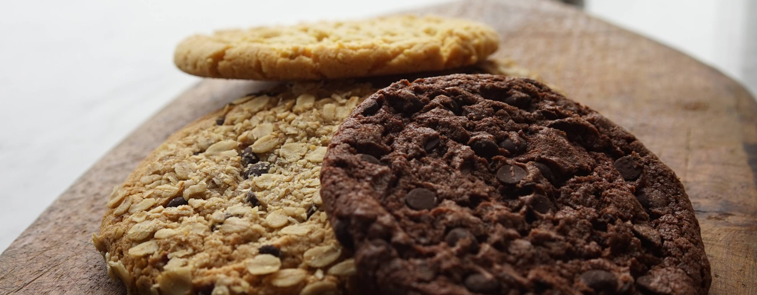 Tre cookies ligger på en skärbräda i trä på ett vitt bord. Två ljusbruna och en mörkbrun.