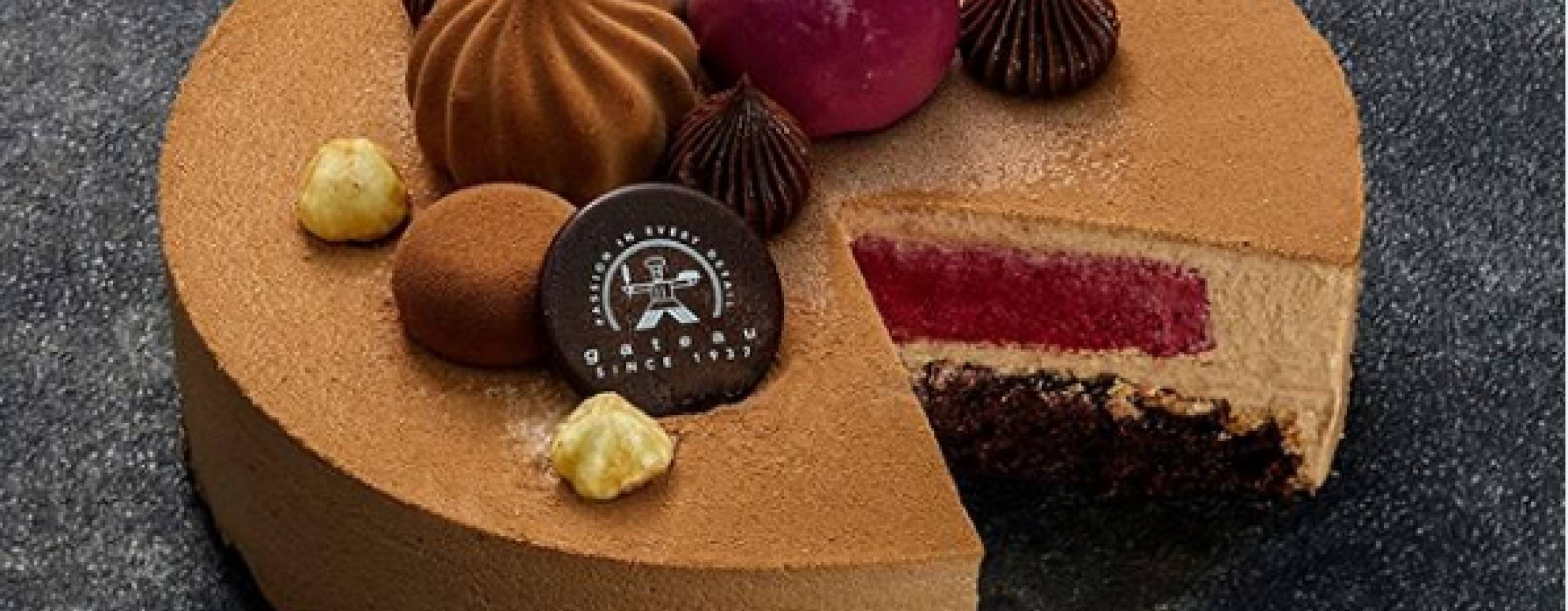 Foto: Gateau En brun rund tårta där en bit har tagits så att innehållet med svartvinbär och mousse syns. Toppat med dekorationer från Gateau