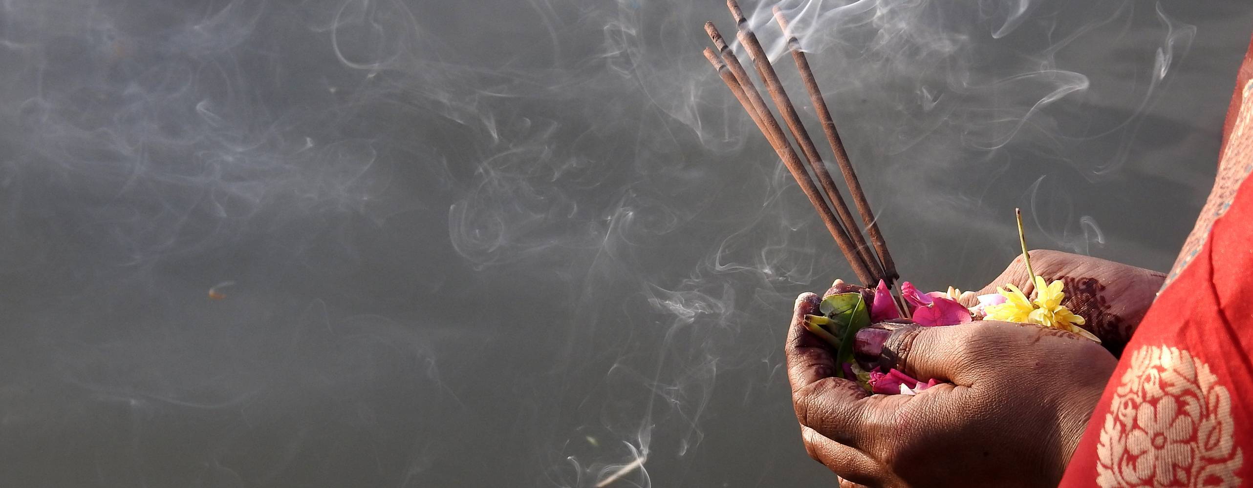 Händer som håller i blommor och rökelser mot grå bakgrund/mörkt vatten