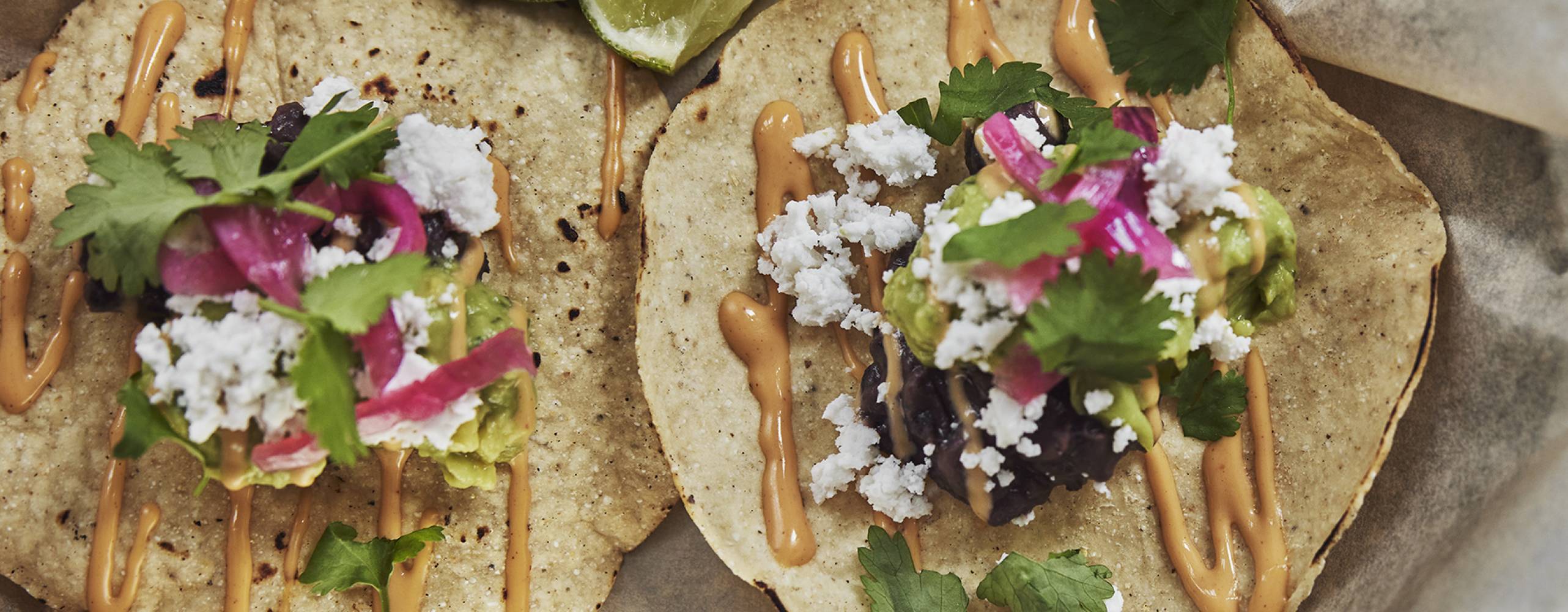 Två, små tacobröd fyllda med bl.a. rödlök, avocado, koriander, vegansk ost och sås, liggandes på bakplåtspapper bredvid limeklyftor.