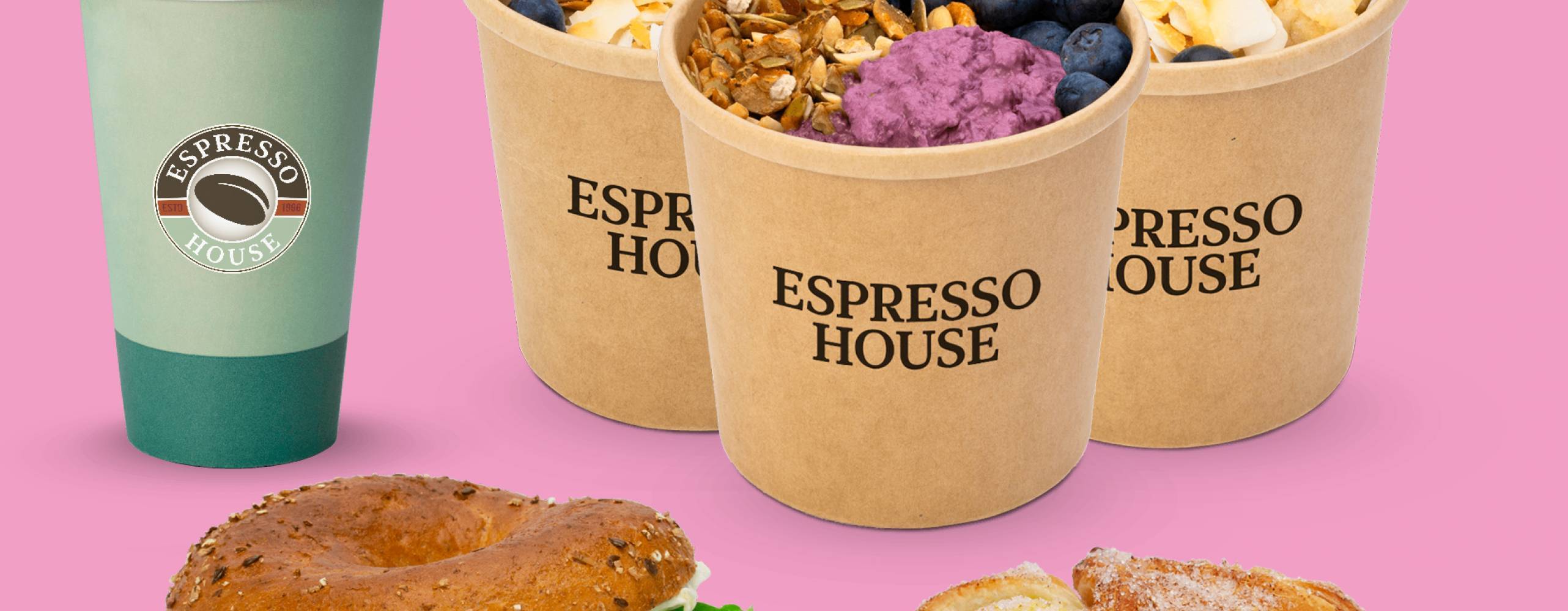 Cafe Latte i pappersmugg, tre små papperskoppar med overnight oats resp. chiapudding med färgglada toppings, avokadobagel och croissant mot rosa bakgrund.