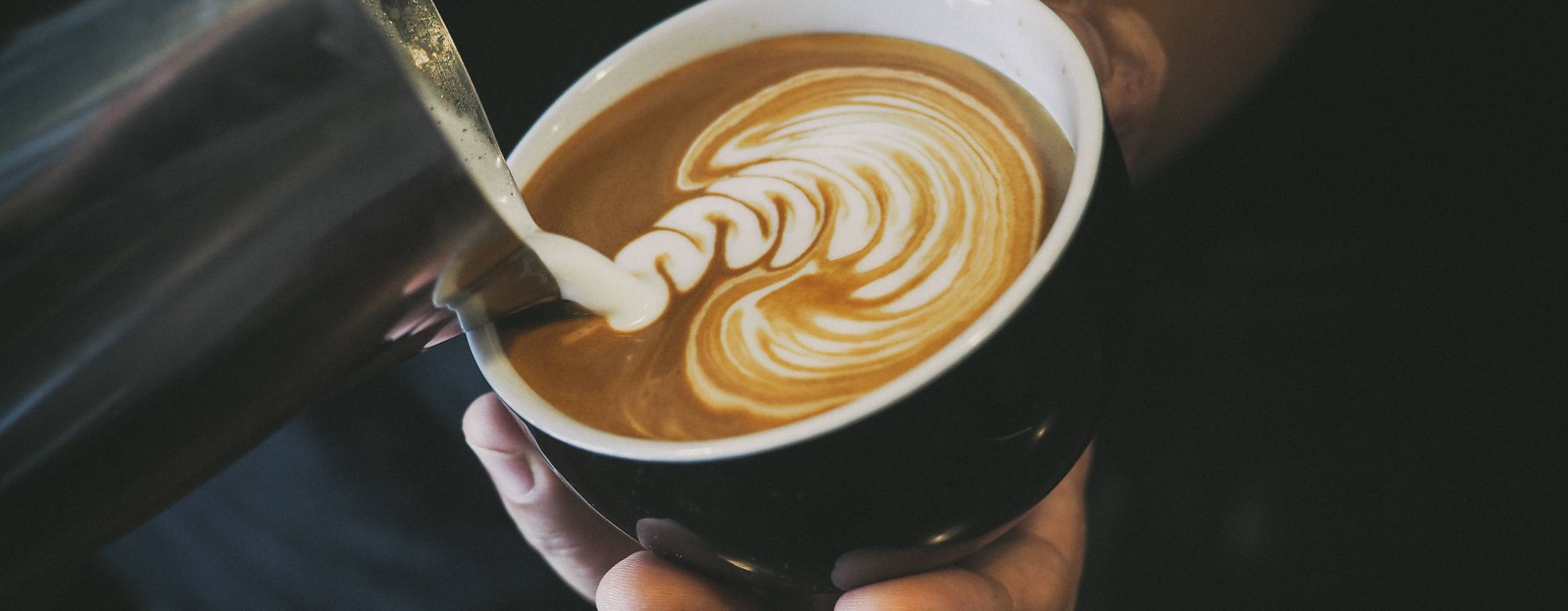 &quot;Mjölk&quot; hälls i kopp med kaffe. Koppen hålls i en hand i dovt belyst miljö.