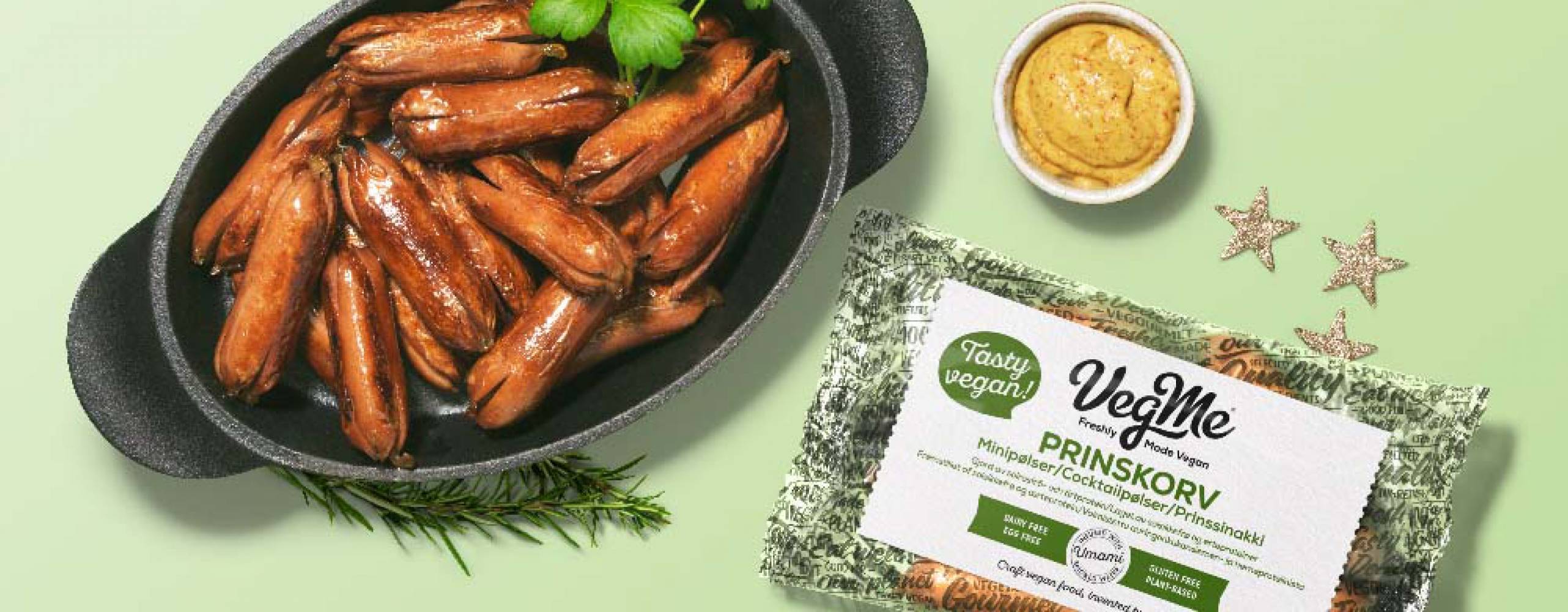 Foto: VegMe Prinskorv från VegMe i gjutjärnsgryta mot grönbakgrund, med förpackning och en liten skål senap bredvid.