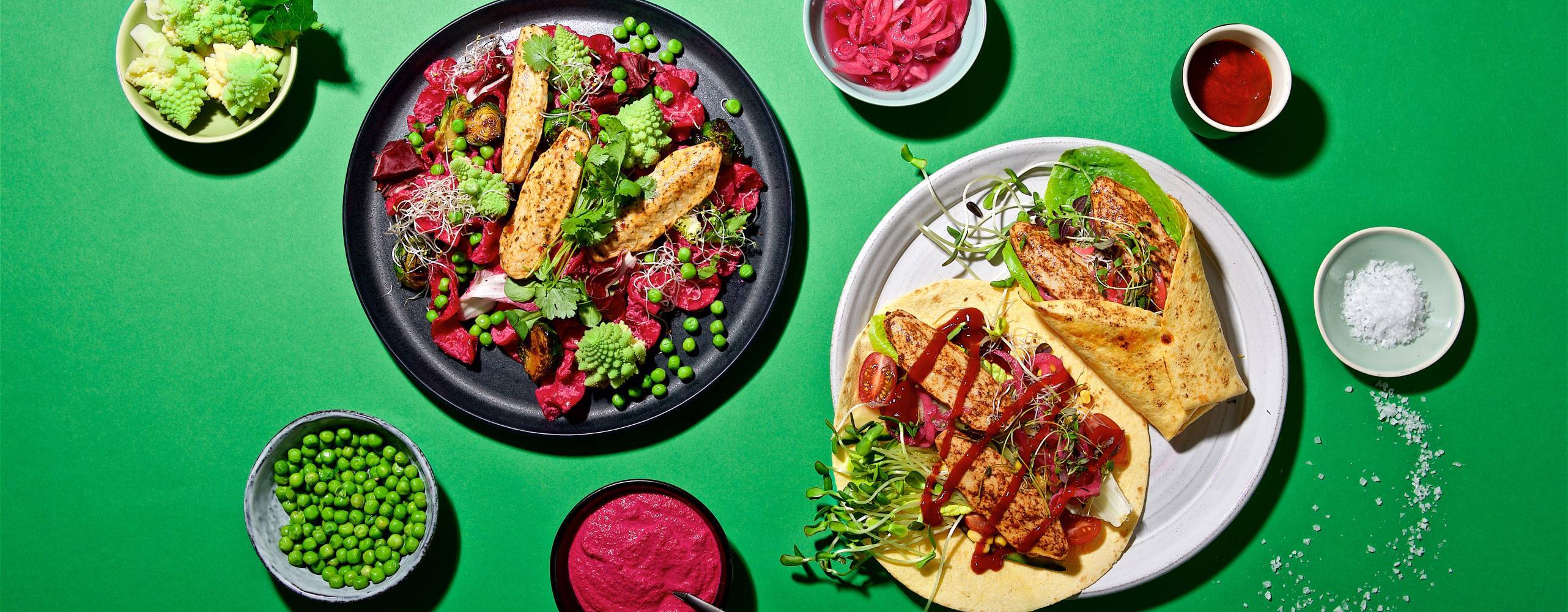 Produktfoto: Quorn veganska filléer i en sallad på en tallrik och en i wrap på en annan tallrik uppdukat på ett grönt bord med tillbehör.
