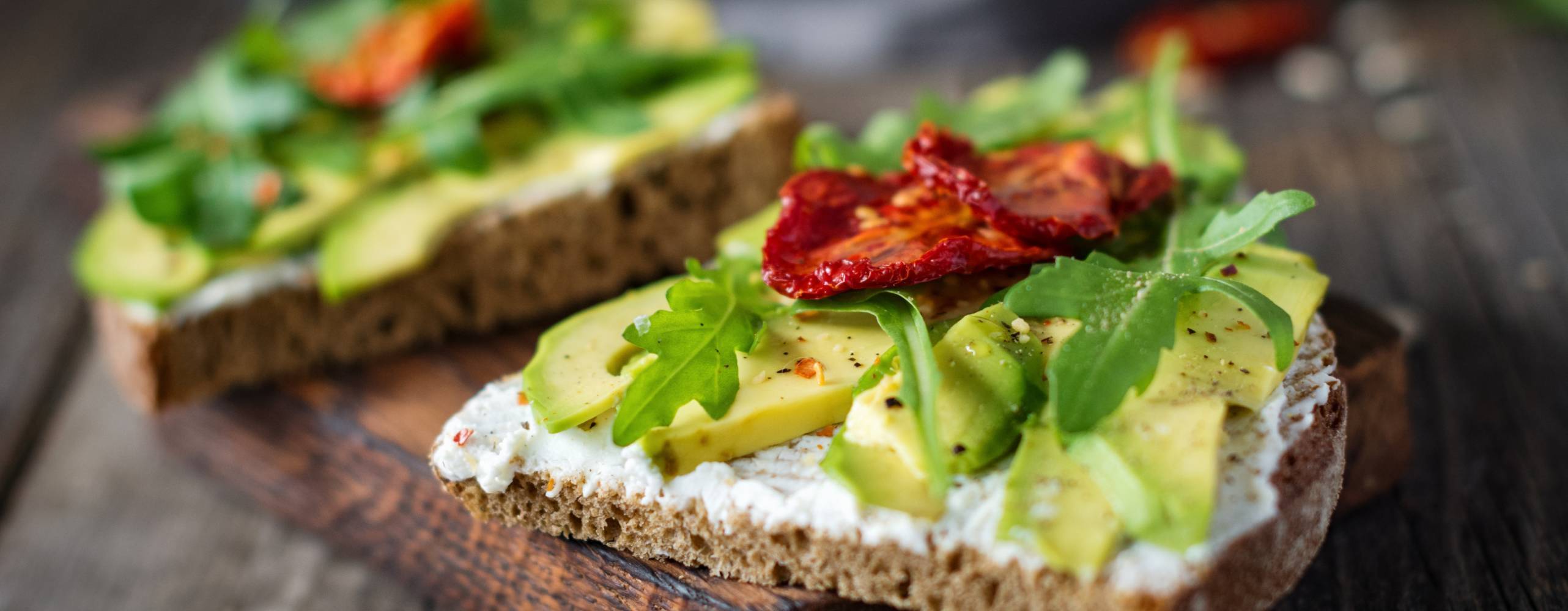 Två rågbrödsmackor med avokado och soltorkade tomater på en skärbräda.