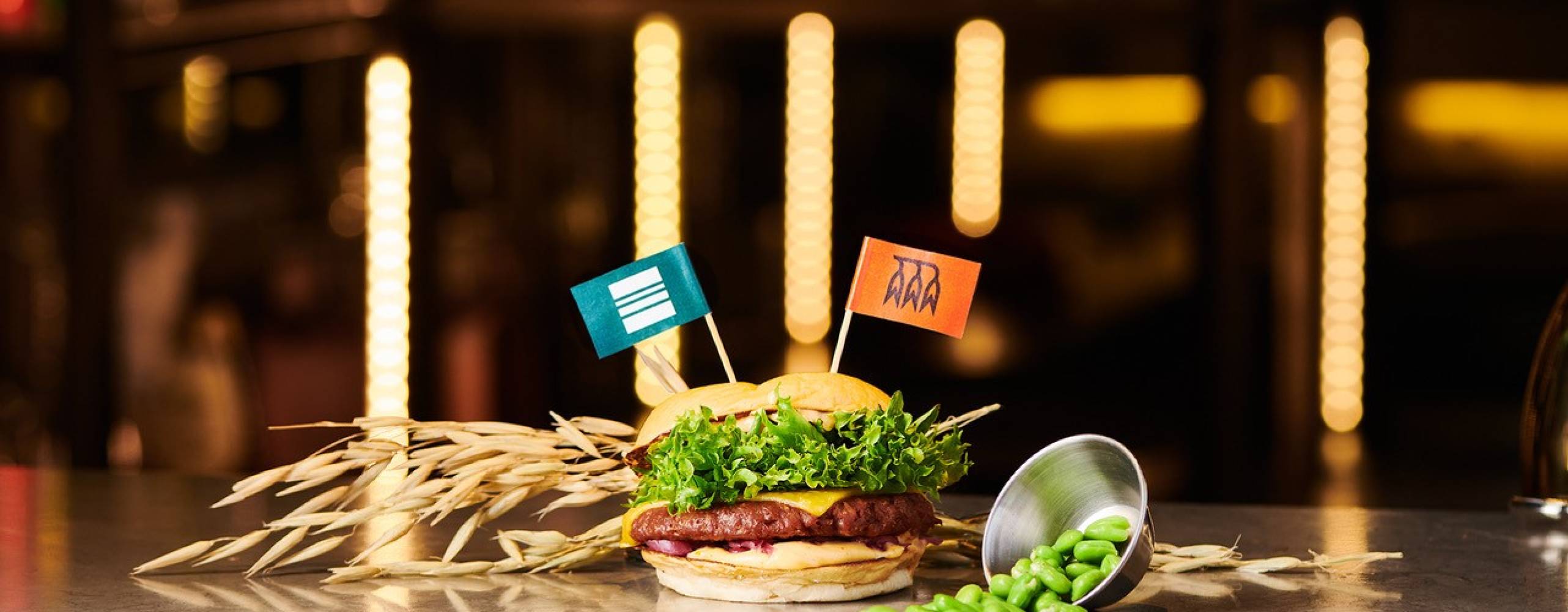Produktfoto: Havredals En växtbaserat burgare på ett bord med två flaggor i brödet dekorerat med havre och sojabönor runt burgaren.