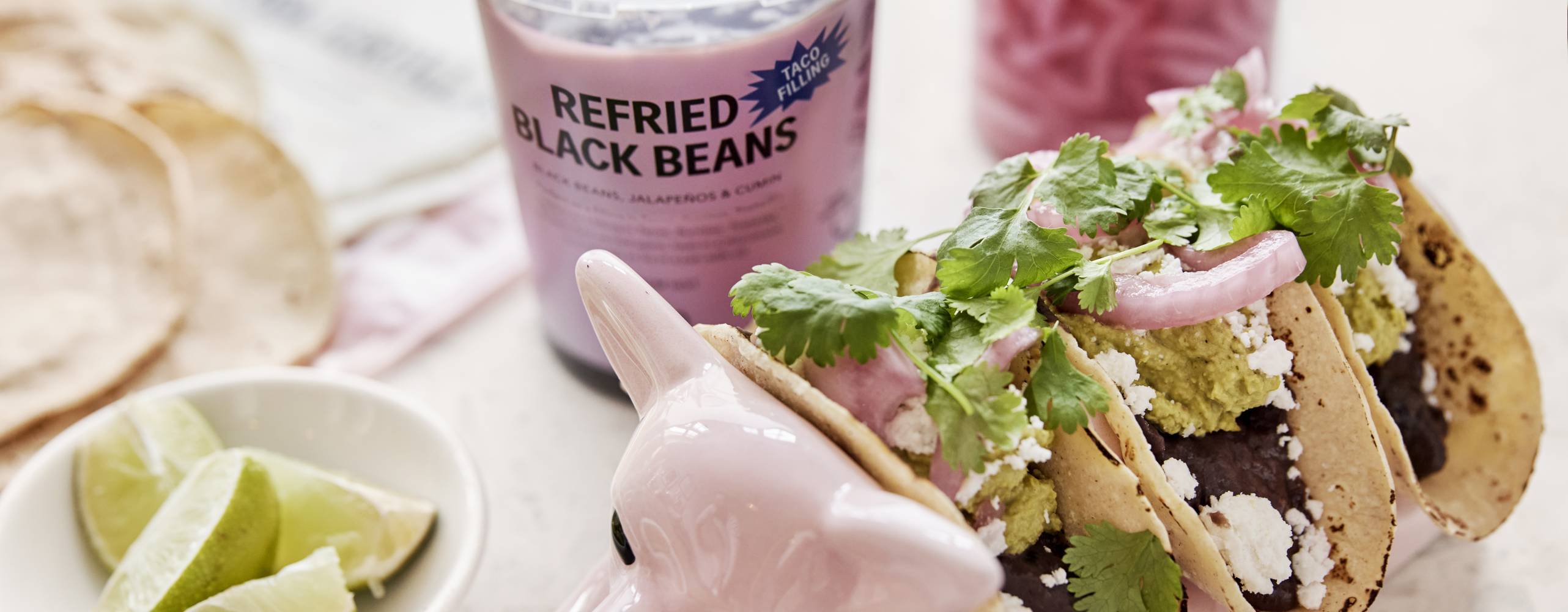 Mjuka fyllda veganska tacos uppställda i en keramikhund med läsk, lime och picklad rödlök på sidan om.