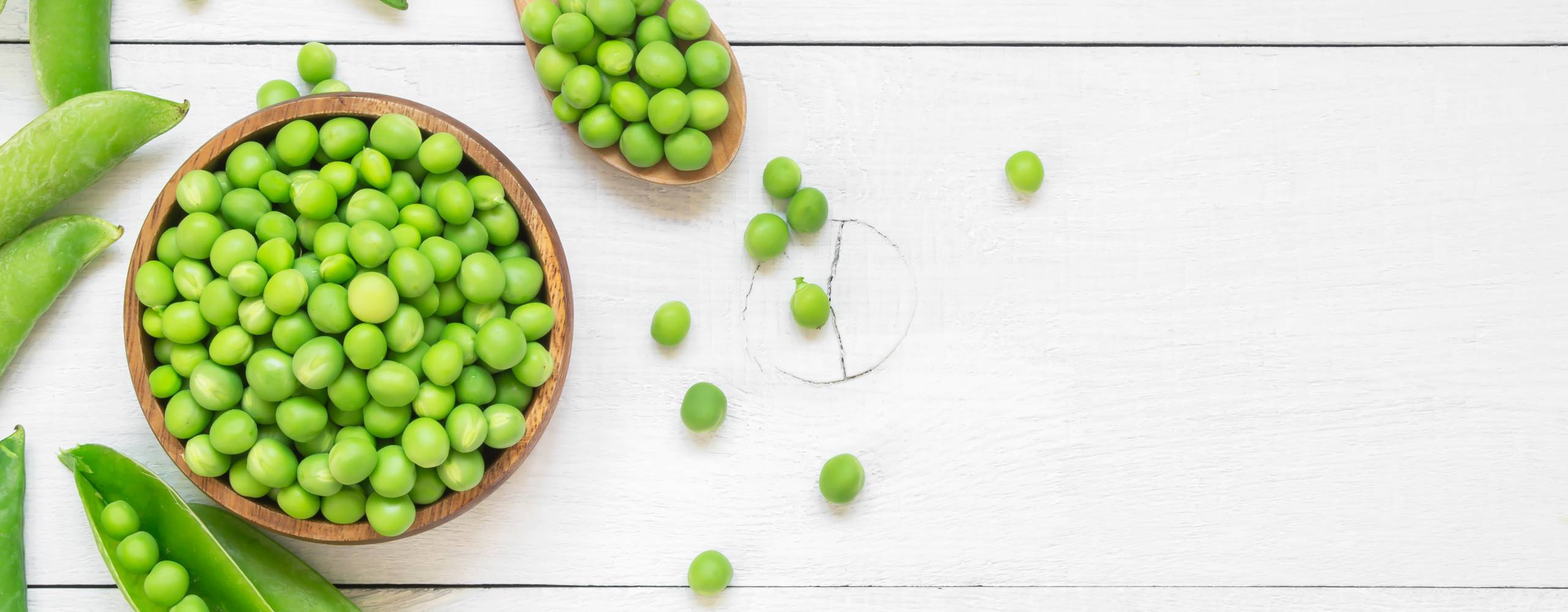 gröna ärtor i en träskål bredvid en träsked och ärtskidor på ett vitt bord.