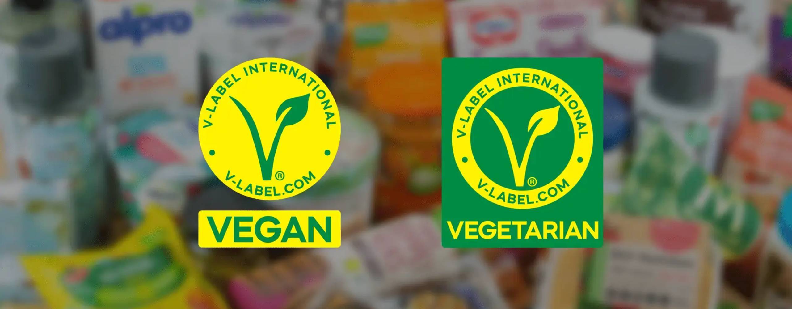 Två uppdaterade V-label loggor mot en suddig bakgrund av livsmedelsprodukter