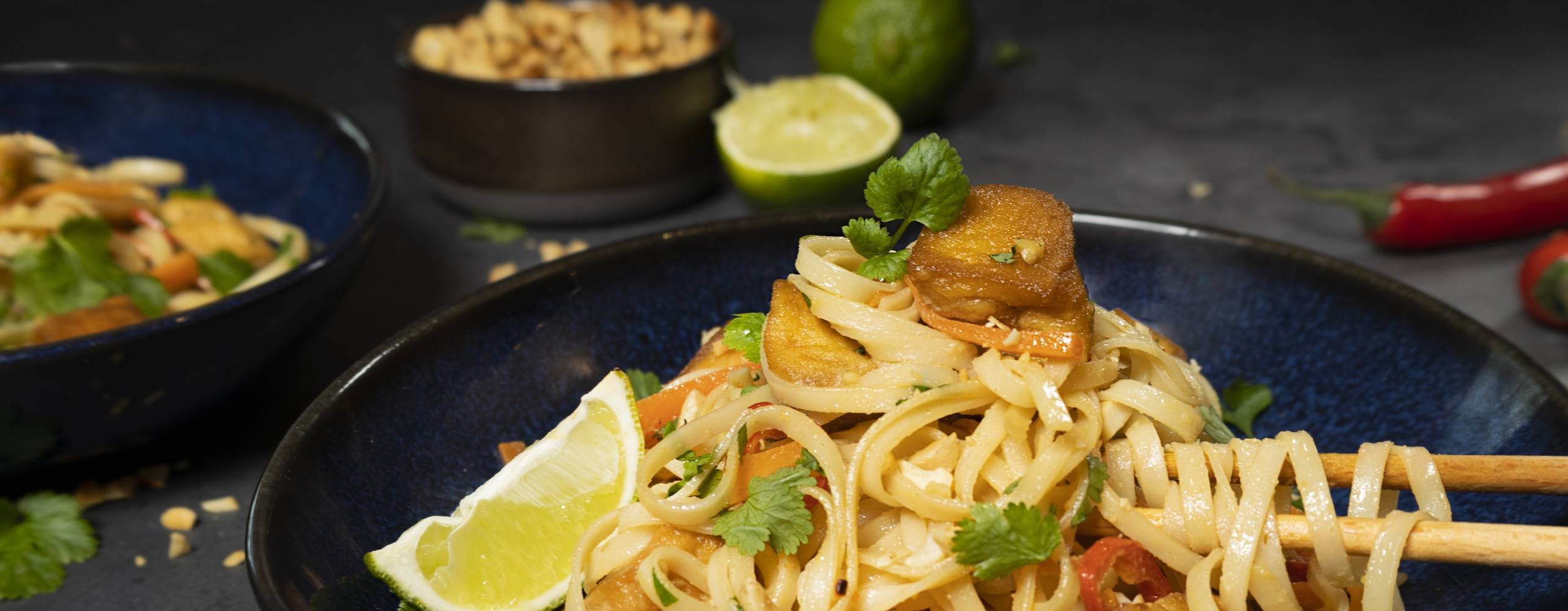Veganska pad thai med tofu i en mörkblå djuptallrik mot en mörkgrå bakgrund.