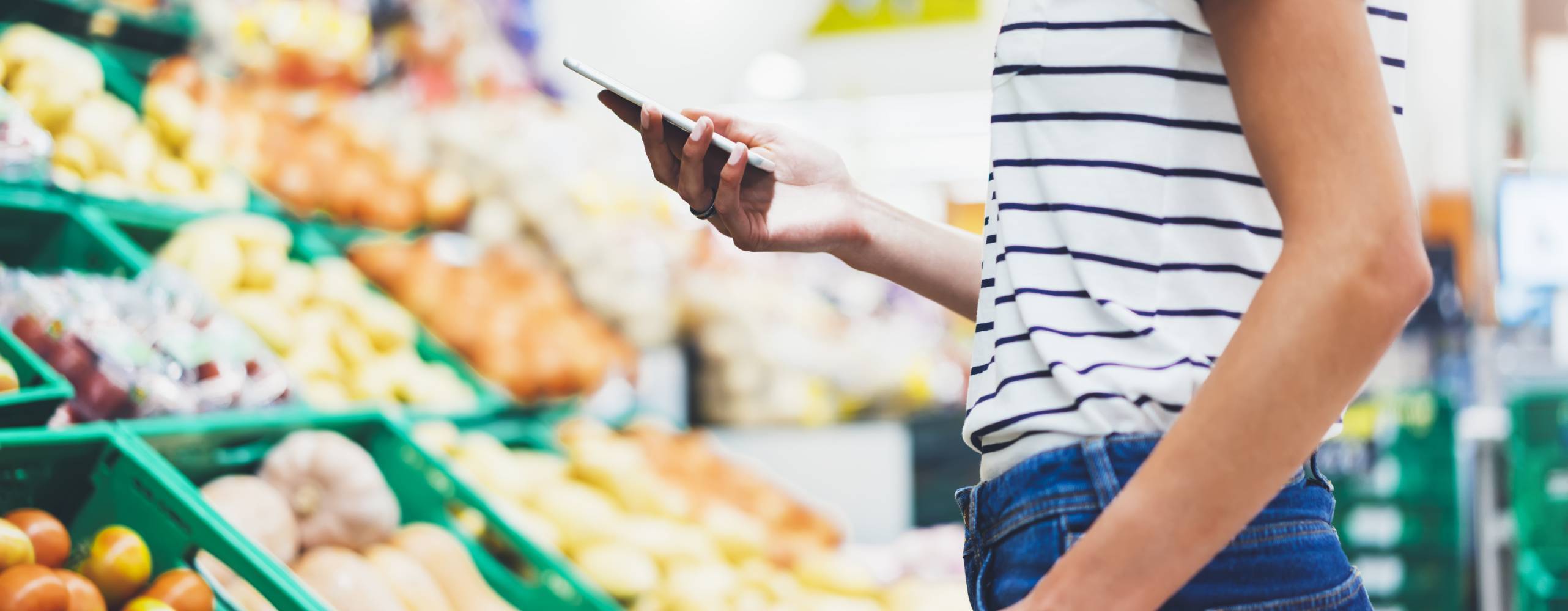 Ung kvinna handlar hälsosam mat i stormarknad suddig bakgrund. Kvinnliga händer köper naturprodukter med smart telefon i butik. Hipster på livsmedelsbutiker med hjälp av mobil. Person som jämför priset på produkter