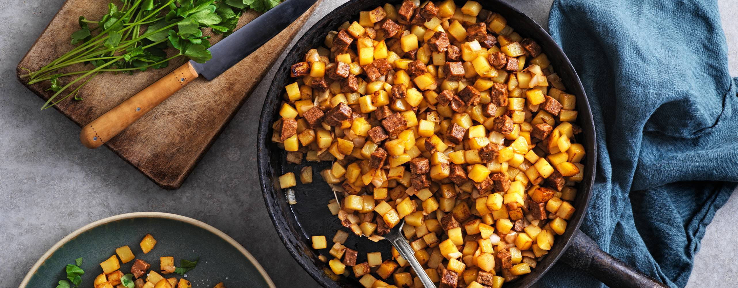 Vegansk pytt i en stekpanna bredvid en tallrik med pytt och en skärbräda med örter