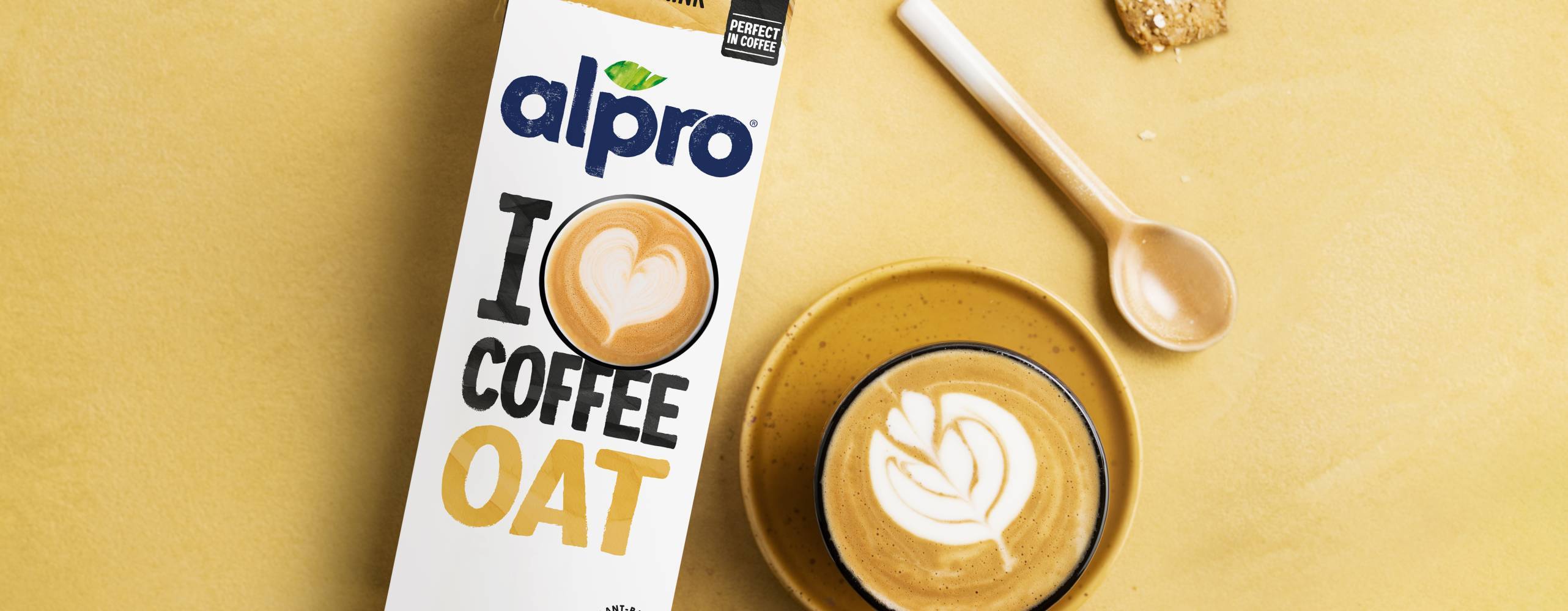 Förpackning med växtbaserad dryck bredvid en kopp latteart på ett gult bord