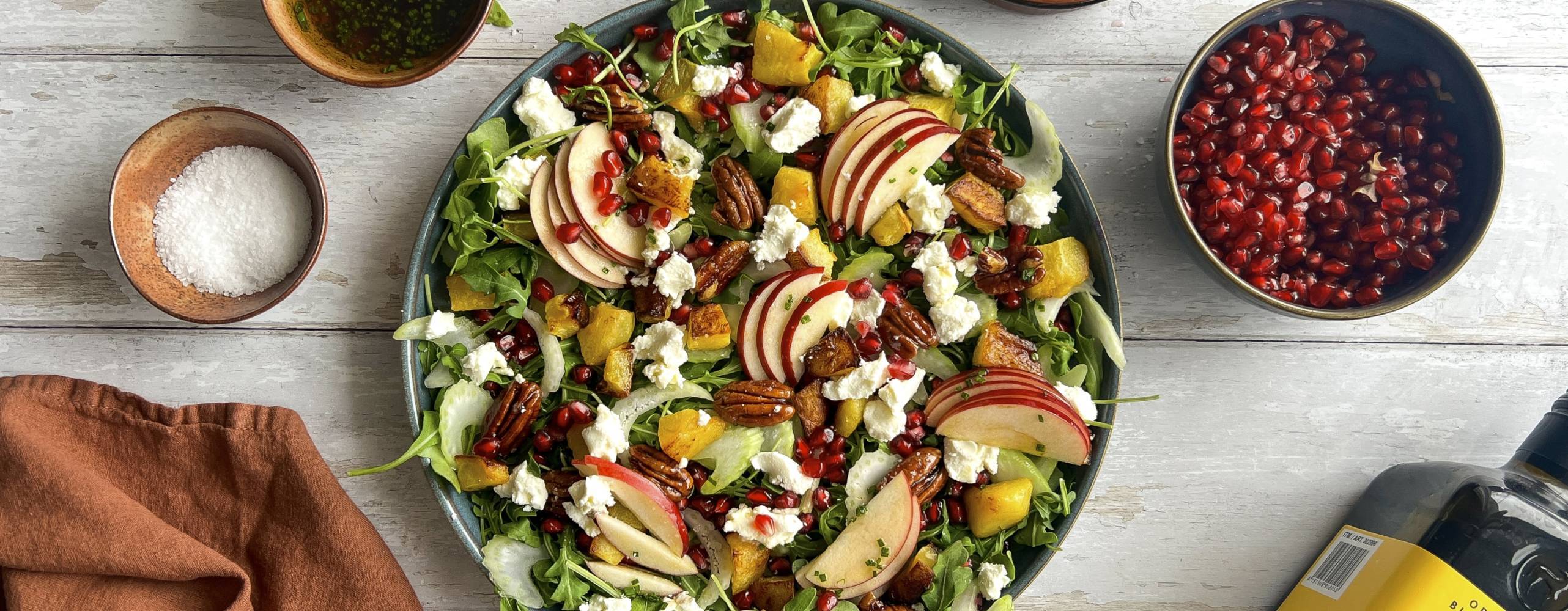 Sallad i en skål på ett vitt bord med granatäpplekärnor, mango, olivolja och en brun kökshandduk runt om tallriken.