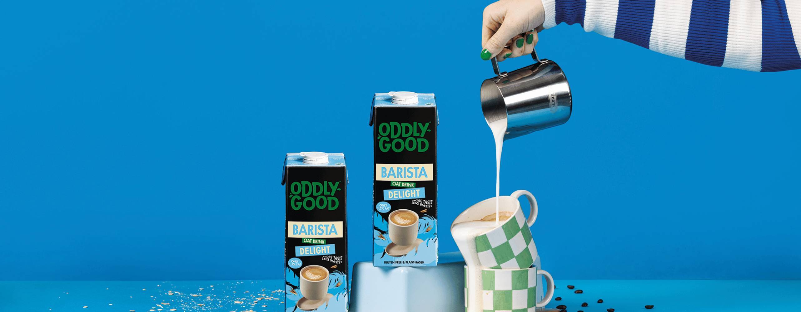 två förpackningar med havredryck och en hand som häller haredryck i en kopp mot en blå bakgrund.