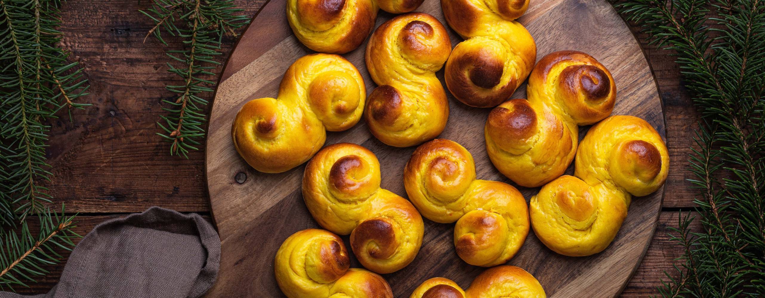 Freshly baked homemade Swedish traditional s-shaped saffron buns, also known as lussebullar or saffransbröd. Seen from above flat lay on a dark wooden table surrounded by spruce branches.