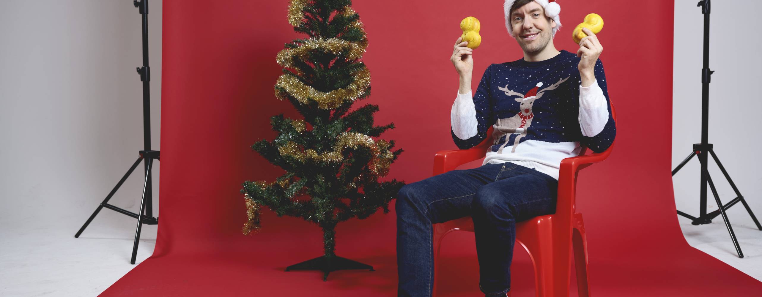 En man som sitter på en stol framför en röd fotovägg. Han håller upp lussebullar, har en jultröja på sig och sitter bredvid en julgran.