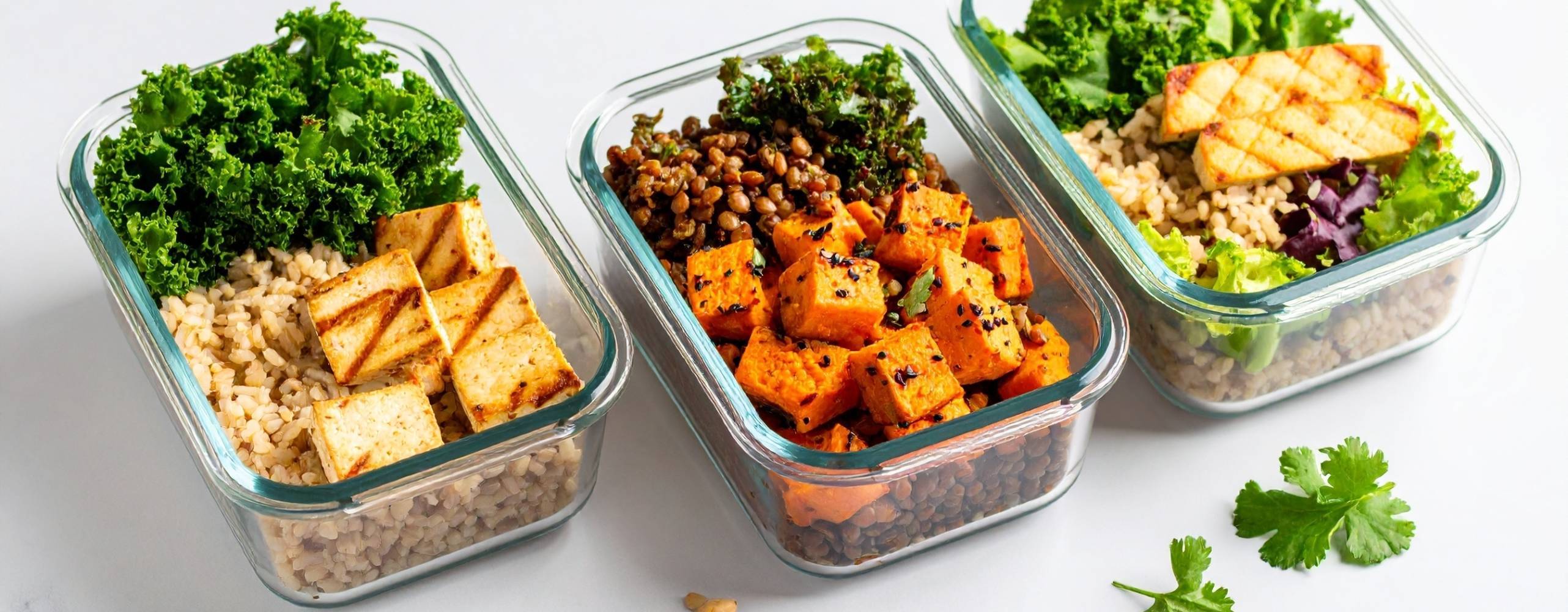 Tre klara glasmåltidsbehållare innehåller nyttiga veganska luncher med råris, grillad tofu, linser, rostad sötpotatis och färska grönsaker.